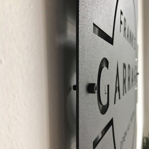 May include: Black metal sign with the text "FRANK'S GARAGE" cut out in a circular shape. The sign is mounted on a white wall.