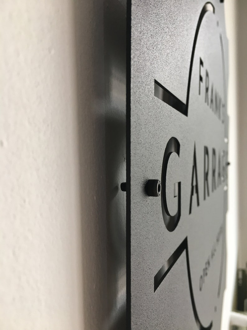 May include: Black metal sign with the text "FRANK'S GARAGE" cut out in a circular shape. The sign is mounted on a white wall.