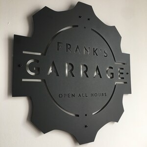 May include: Black metal sign with a gear shape and the text "Frank's Garage Open All Hours" in a circle.