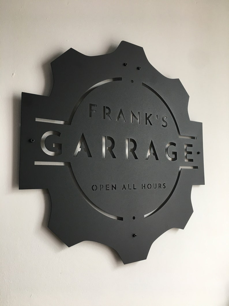 May include: Black metal sign with a gear shape and the text "Frank's Garage Open All Hours" in a circle.
