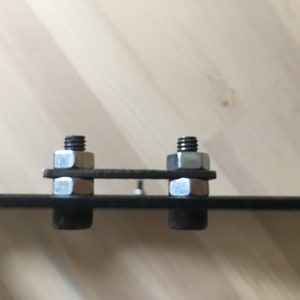 May include: Two silver nuts and bolts are attached to a black metal bar. The nuts and bolts are secured with a washer on each side.
