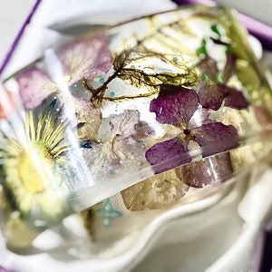 Peut inclure: Un bracelet en résine transparente avec des fleurs pressées et des paillettes. Les fleurs sont violettes, roses et jaunes.