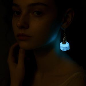 以下が含まれることがあります： 銀色のチェーンに吊るされた、光る正方形のイヤリング。イヤリングは柔らかい青色の光を放ち、暗い背景とのコントラストを生み出しています。画像はイヤリングとそれを身に着けている人に焦点を当てています。
