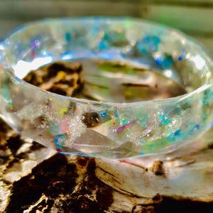 May include: A clear resin bangle bracelet with embedded iridescent glitter and colourful accents. The bracelet is round and sits on a piece of wood. The glitter includes shades of blue, green, pink, and silver.