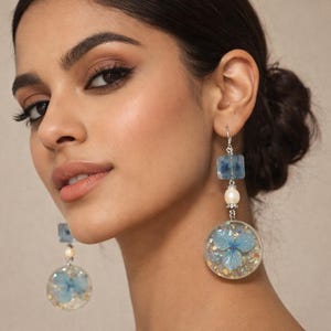 Puede incluir: Pendientes colgantes con una pieza superior cuadrada de resina azul, una pequeña perla blanca y un gran colgante circular de resina que contiene una flor azul y purpurina. Los pendientes cuelgan de ganchos y piezas de metal plateado.