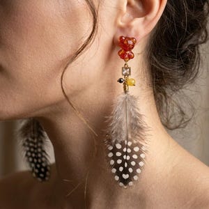 Puede incluir: Primer plano de una oreja adornada con un pendiente único. El pendiente presenta un elemento rojo en forma de oso, un cristal cuadrado y una pluma gris con puntos blancos. Un pendiente llamativo.