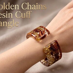 Könnte beinhalten: Nahaufnahme eines Resin-Manschettenarmbands mit goldenen Kettenakzenten. Das transparente Armband hat ein braunes und goldenes Design. Der Text "Golden Chains Resin Cuff Bangle" ist sichtbar.