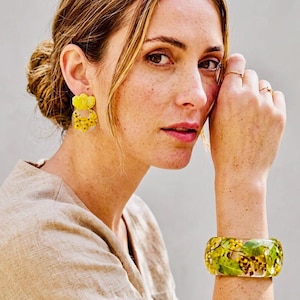 Puede incluir: Un par de pendientes de resina amarilla con motivos florales y una pulsera rígida a juego de resina transparente que contiene hojas verdes y pequeñas flores amarillas incrustadas. Los pendientes tienen una parte superior amarilla.