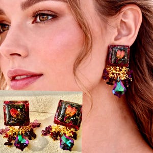 May include: Square resin earrings with a dried flower encased in the center. The earrings have a dark red border, gold filigree, and a large, iridescent, teardrop-shaped crystal. The earrings are dangling.