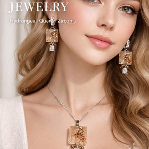 Puede incluir: Juego de joyas con collar y pendientes. Las joyas tienen un colgante y pendientes cuadrados con flores secas encapsuladas en resina transparente, con detalles de cuarzo y circonita. El texto "Real Flower Jewelry" es visible.