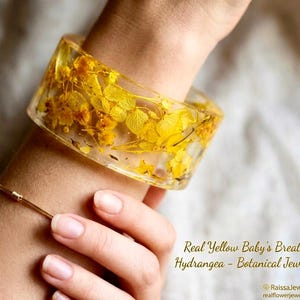 Könnte beinhalten: Ein klarer Harz-Armreif, gefüllt mit gelben Schleierkraut- und Hortensienblüten. Der botanische Schmuck wird am Handgelenk getragen, darunter ein zartes goldenes Armband. Der Text lautet "Real Yellow Baby's Breath & Hydrangea - Botanical Jewelry."