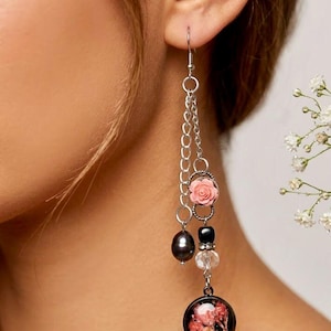 Puede incluir: Pendientes de cadena plateada con una rosa rosa, perlas negras y una cuenta transparente. Los pendientes tienen un colgante redondo con flores secas. El texto "baby breath black river pearls" está en la parte inferior.