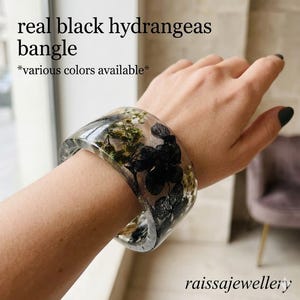Könnte beinhalten: Ein klarer Harzarmreif mit echten schwarzen Hortensien und anderen getrockneten Blumen. Der Armreif wird am Handgelenk getragen. Der Text auf dem Bild lautet "real black hydrangeas bangle" und "*various colors available*". Der Markenname "raissajewellery" ist ebenfalls sichtbar.
