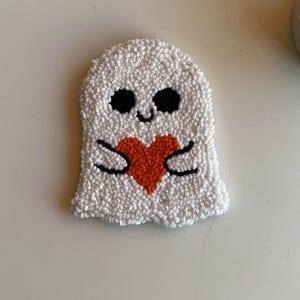 May include: A white punch needle ghost coaster with a textured, looped yarn surface. The ghost features two round black eyes, a small black smile, and two black arms holding a bright orange heart centered on its body.