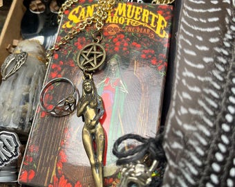 May include: An assortment of occult items, including a tarot deck, a metal pendant of a nude figure, a pentagram charm, and a silver ring. The tarot deck is labelled "Santa Muerte Tarot of the Dead". The image has a dark, mystical aesthetic.