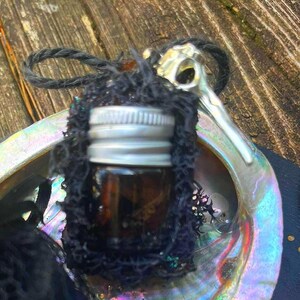 May include: A small, brown glass bottle with a silver cap, wrapped in black netting, sits inside an iridescent abalone shell. A black cord is attached to the bottle. The shell is resting on a wooden surface.