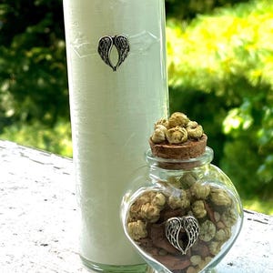 May include: A tall, pale green candle with a silver wing charm and a heart-shaped glass jar filled with dried flowers and a cork stopper. The jar also has a silver wing charm. The background is blurred green foliage.