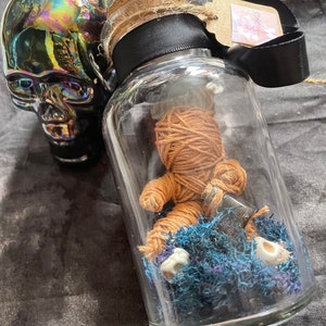 May include: A clear glass bottle with a cork stopper, black ribbon, and a small tag. Inside, a brown yarn doll rests on blue and purple moss, with small white skulls. A shiny, iridescent skull is next to the bottle.