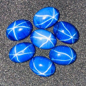 Puede incluir: Seis piedras preciosas azules de forma ovalada dispuestas en un grupo. Cada piedra lisa y pulida presenta un distintivo patrón de estrella blanca de seis puntas en su superficie, creando un efecto luminoso sobre el color azul profundo de las piedras talladas en cabujón.