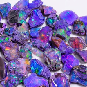 Puede incluir: Una colección de piedras de ópalo en bruto e irregulares con un color base púrpura vibrante. Cada piedra muestra un juego de colores único con destellos de verde neón, rojo brillante y azul eléctrico que se reflejan en las superficies cristalinas.