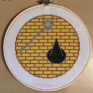 May include: A yellow brick road with a black witch's shoe and a silver wand, embroidered on a white canvas in a wooden embroidery hoop.