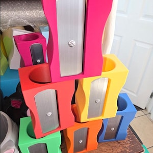 Könnte beinhalten: Ein Stapel bunter Anspitzer in Pink, Rot, Gelb, Grün, Orange und Blau. Jeder Anspitzer hat eine silberne Klinge und eine zylindrische Öffnung für Bleistifte. Die Anspitzer sind in einer ansprechenden, gestapelten Anordnung angeordnet.