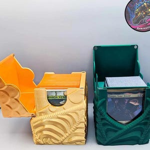 Könnte beinhalten: Zwei dekorative Kartenaufbewahrungsboxen. Eine ist goldfarben mit offenem Deckel, die andere grün mit offenem Deckel. Beide Boxen haben ein strukturiertes Design. Die goldene Box enthält orangefarbene Karten, und die grüne Box enthält Karten mit Text.