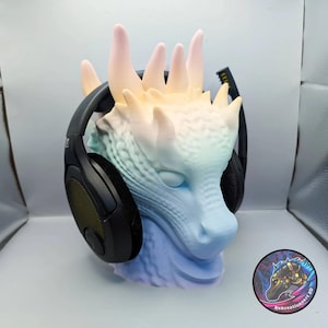 May include: A dragon head sculpture featuring a pastel gradient of blue, green, and pink, serving as a stand for black over-ear headphones. The sculpture has textured scales and multiple horns. A circular logo with the text Kekreationsart 3D is visible.