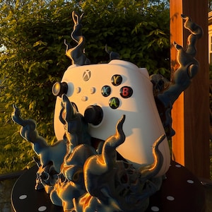 Può includere: Un controller Xbox bianco è posizionato su un supporto stampato in 3D a forma di fiamme. Il supporto è di colore blu scuro con un riflesso metallico. Il controller è posizionato su una base circolare nera.