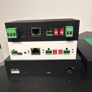 May include: A stack of black and white electronic devices featuring various ports, switches, and green connectors. The rectangular devices likely serve networking or control functions. Red switches are labelled "ON".