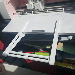 Può includere: Una macchina da taglio Cricut bianca con interno rosa. La macchina ha un vassoio bianco e una struttura di supporto bianca. Il logo Cricut è visibile sulla parte superiore della macchina.