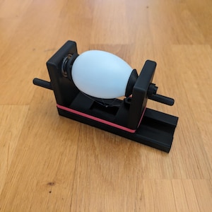 May include: A black plastic device with a white egg-shaped object inside. The device has a rubber band and a small handle.