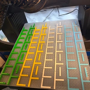May include: A clear plastic board with a grid of holes, featuring colorful plastic pieces in green, yellow, white, and blue. The pieces are arranged in a pattern, creating a visual puzzle or game. The board is set against a dark background.
