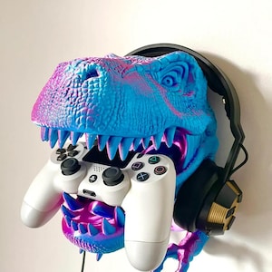 May include: A blue and pink dinosaur head wall mount holding a white PlayStation controller. Black headphones are on the dinosaur's head. The dinosaur has sharp teeth and is mounted on a white wall.