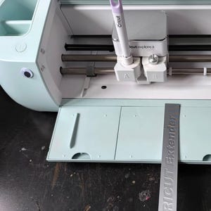 May include: A mint green Cricut Explore 3 machine with a white and purple Cricut pen. The machine's cutting head is visible, along with a silver Cricut Extender. The machine is open, revealing its interior components.
