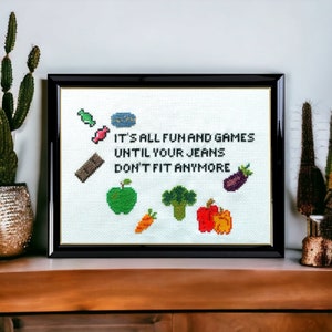 May include: A framed cross-stitch with the text "It's all fun and games until your jeans don't fit anymore." The cross-stitch features a variety of colorful foods, including a green apple, a carrot, a broccoli, a red and orange pepper, and a purple eggplant.