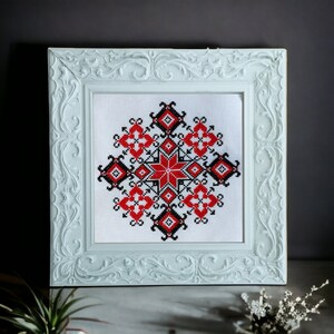May include: A white framed cross-stitch embroidery with a red, black, and white geometric pattern. The pattern is a stylized star with a repeating design.