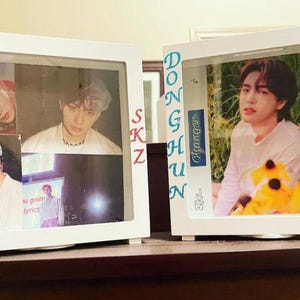 May include: Two white framed shadow boxes with photos of people. One box has the text "BANGCHAN" in red and the other has the text "DONGHUN" in blue. The boxes are on a dark surface.