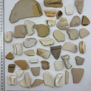 May include: A collection of pottery shards in beige, tan, and brown hues, arranged on a white surface. The fragments vary in size and shape, with some showing remnants of patterns or textures. A ruler is placed along the bottom edge.