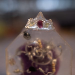 Puede incluir: Una pirámide de resina transparente con un objeto morado y plateado en su interior. La parte superior de la pirámide tiene una pequeña corona con una gema rosa, verde y amarilla.