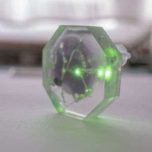 Op de afbeelding: Een transparante, achthoekige harshanger met een klein printplaatje en twee groene LED-lampjes erin. De hanger ligt op een wit oppervlak.