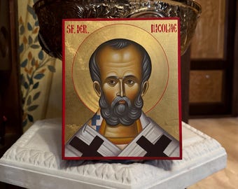 May include: A hand-painted icon of Saint Nicholas, also known as Santa Claus, with a gold background and a red border. The icon is framed in a wooden frame and has the text "SF. IGR." and "NICOLAE" written on it.