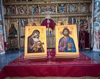 May include: Two religious icons on a red draped table in a church. The icons depict figures with gold backgrounds and are framed in gold. The background features ornate gold detailing and religious artwork.