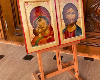 Puede incluir: Dos iconos religiosos enmarcados en un caballete de madera. Los iconos están pintados en un estilo tradicional con detalles en pan de oro. El icono de la izquierda representa a la Virgen María y el Niño, mientras que el icono de la derecha representa a Jesucristo.