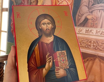 May include: A hand holds a wooden icon of Jesus Christ. The icon is painted in a traditional style with gold accents. The background of the icon is gold with a red border. The icon is surrounded by a red frame.