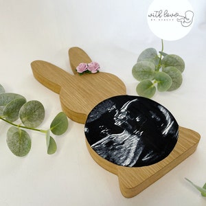 May include: Wooden bunny rabbit cutout with a pink flower embellishment and a black and white ultrasound image in the centre. The text "with love by stacey" is on a white circle in the top right corner.