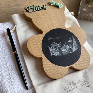 May include: A light-coloured wooden bear-shaped plaque featuring a circular black centre with a white ultrasound image. The name Elliot is in green script, and three small green flowers are attached. A black pen rests on a white fabric drawstring bag.