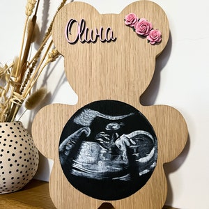 Puede incluir: Placa de madera con forma de oso con una imagen de ultrasonido en blanco y negro en el centro. El nombre "Olivia" está escrito en letras rosas sobre la imagen, y tres flores rosas están colocadas en el lado derecho de la placa.