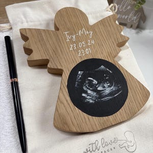 May include: A light-coloured wooden angel-shaped plaque featuring a black circular centre with a white ultrasound image. The wood is engraved with the name Ivy-May, the date 23.05.24, and the time 23:01. A black pen rests on a white fabric bag.