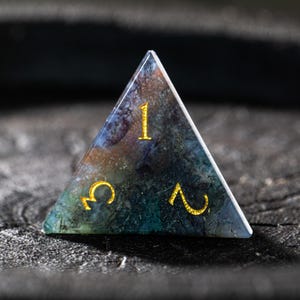Pu&ograve; includere: Un dado triangolare a forma di gemma, con numeri dorati 1, 2 e un simbolo. Il dado ha un aspetto marmorizzato con sfumature di blu, verde e marrone. Un accessorio di gioco unico.